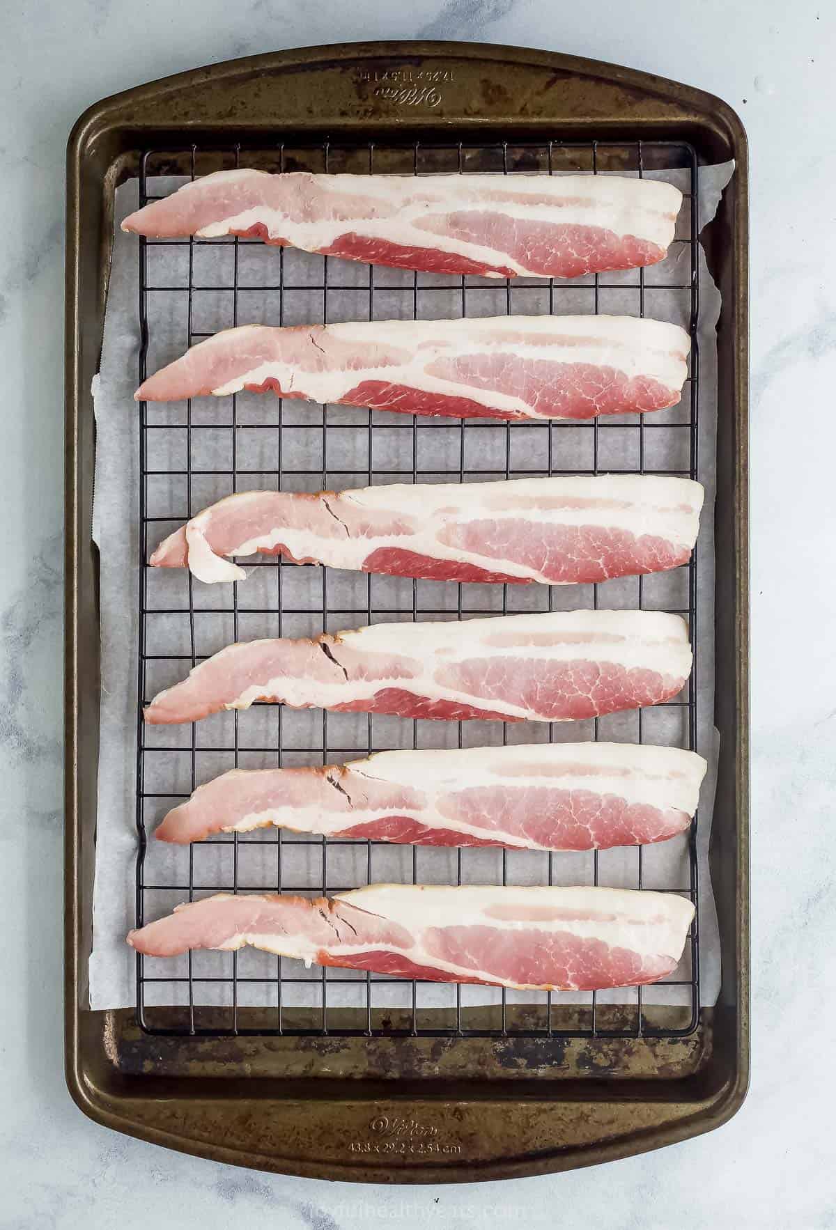 Six strips of bacon sitting on a wire rack positioned on top of a large metal baking sheet