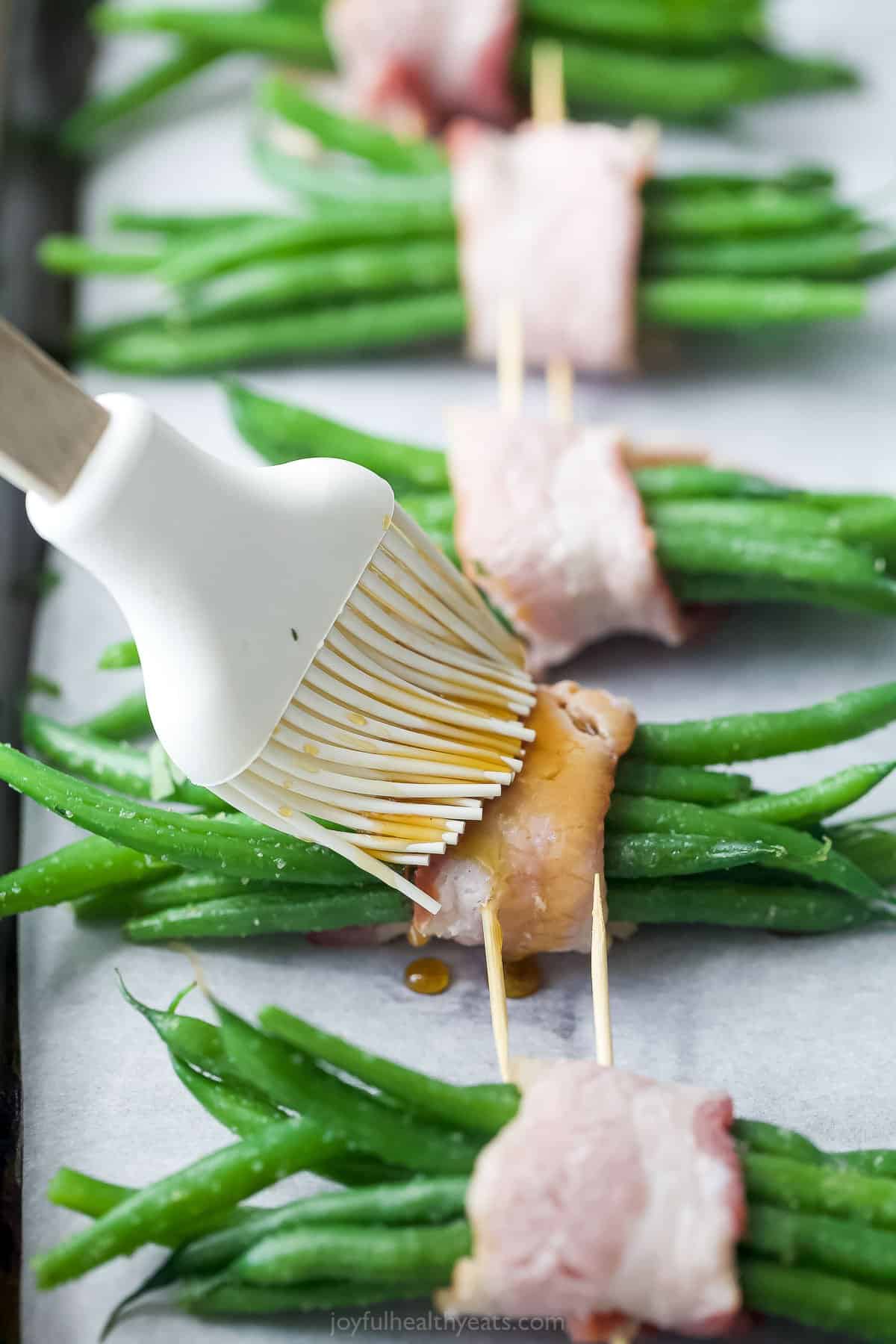 A close-up shot of bacon-wrapped green bean bundles being brushed with maple syrup