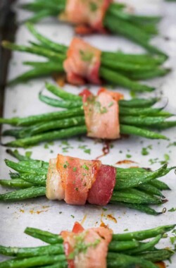 A row of bacon-wrapped green beans on a lined baking sheet with finely chopped parsley sprinkled on top