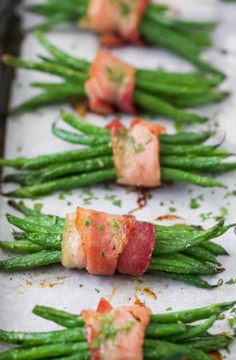 A row of bacon-wrapped green beans on a lined baking sheet with finely chopped parsley sprinkled on top