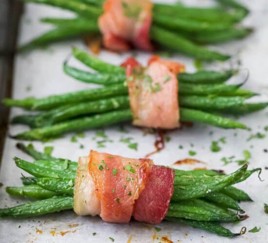 A row of bacon-wrapped green beans on a lined baking sheet with finely chopped parsley sprinkled on top