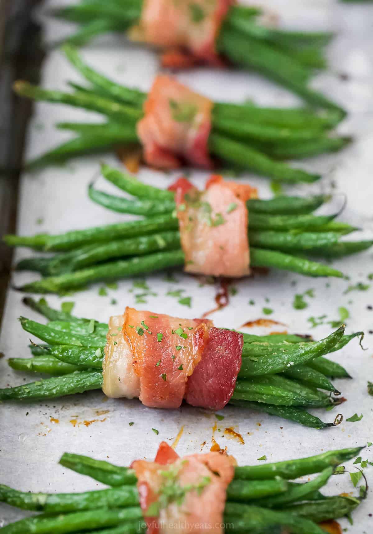 A row of bacon-wrapped green beans on a lined baking sheet with finely chopped parsley sprinkled on top