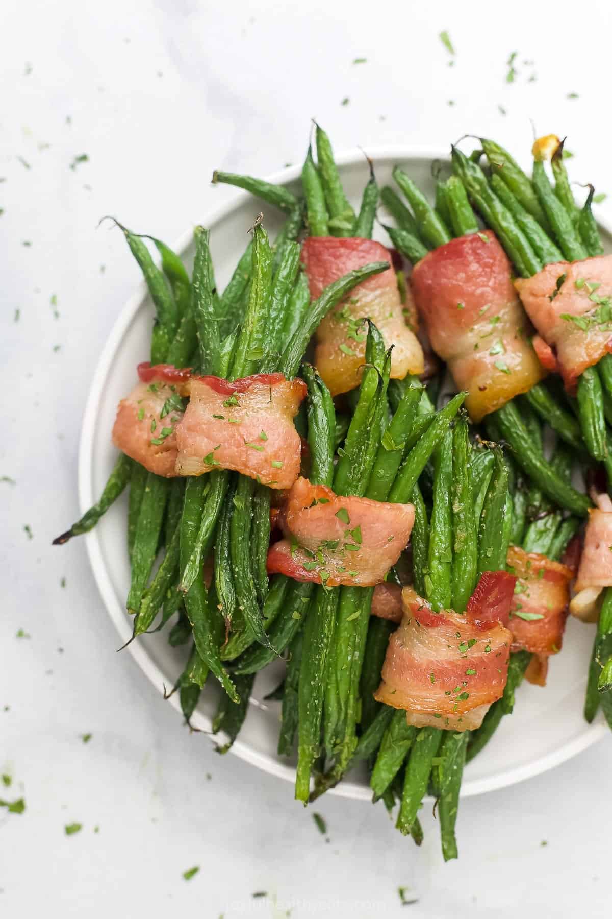 A bunch of bacon-wrapped green bean bundles on a plate with chopped parsley sprinkled about
