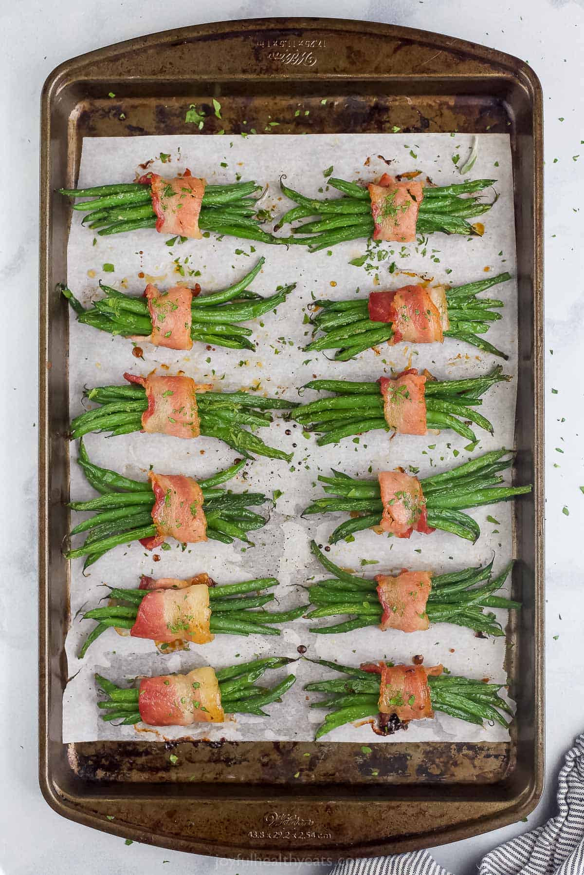 A full batch of green bean bundles on a parchment-lined baking sheet with chopped parsley sprinkled overtop