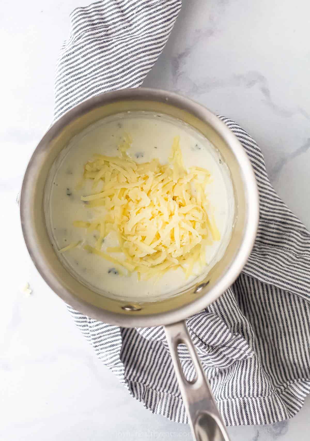 Shredded cheese added to cream sauce in a saucepan.