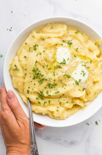 A hand holding a large bowl of parsnip puree topped with butter and chopped parsley