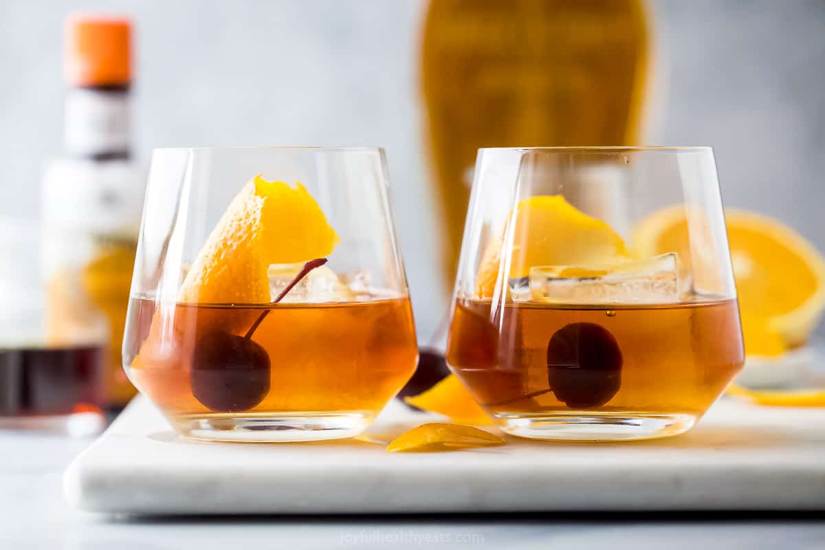 Two maple bourbon cocktails in two glasses sitting side by side on a cutting board