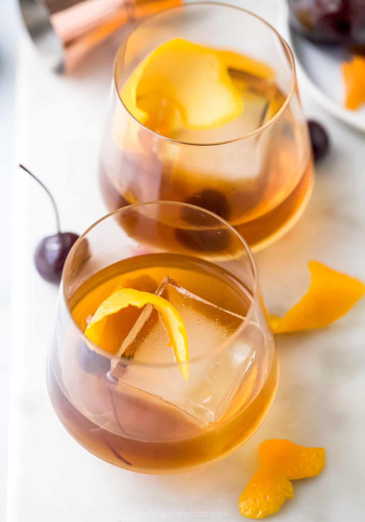 Two maple old fashioned cocktails on a countertop with two cocktail cherries and two small strips of orange peel