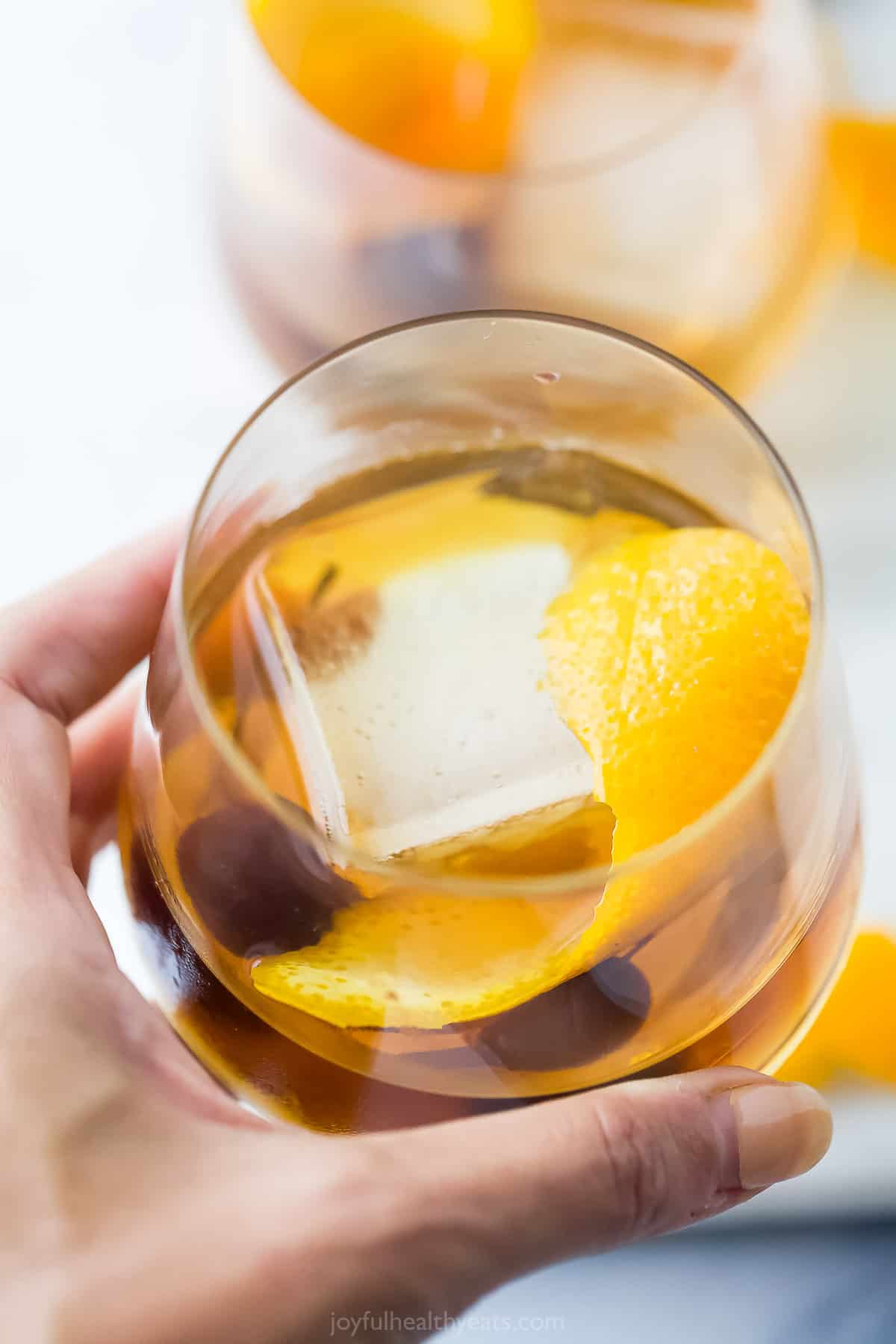 A hand holding a maple old fashioned in a tumbler glass with an ice cube