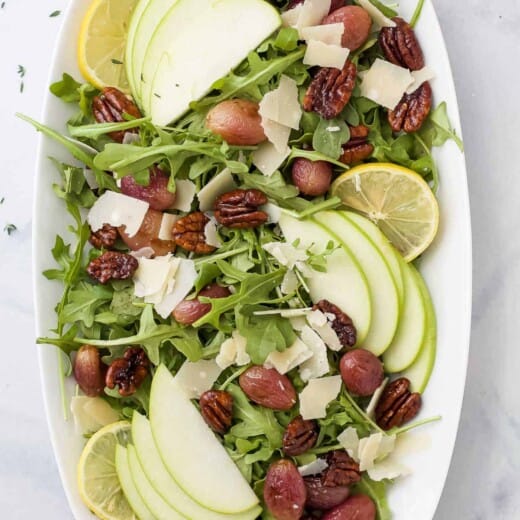 A large serving platter containing a fall salad with candied pecans, apple slices and roasted grapes