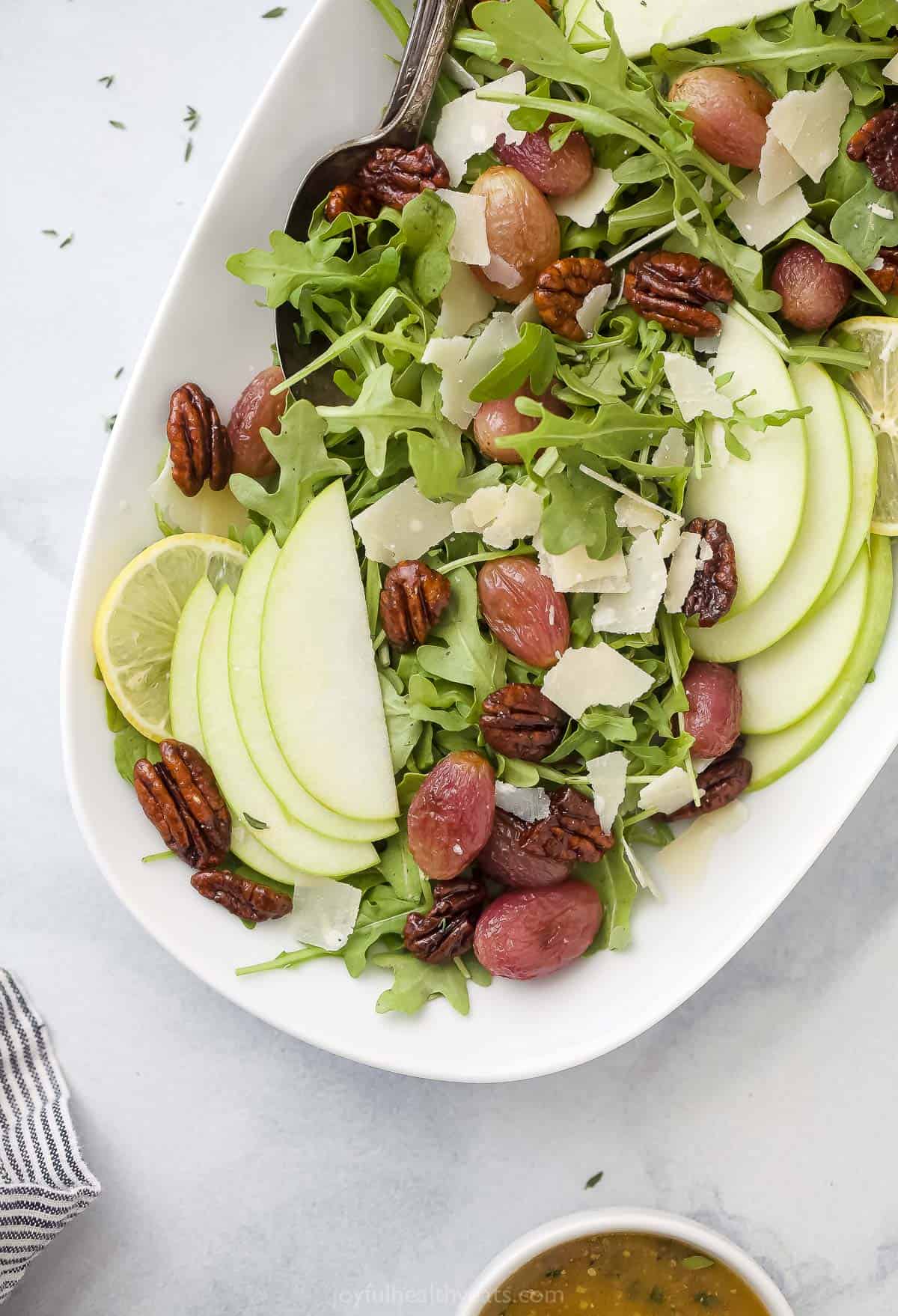 A fall salad with roasted grapes in a large serving platter beside a bowl of dressing and a kitchen towel