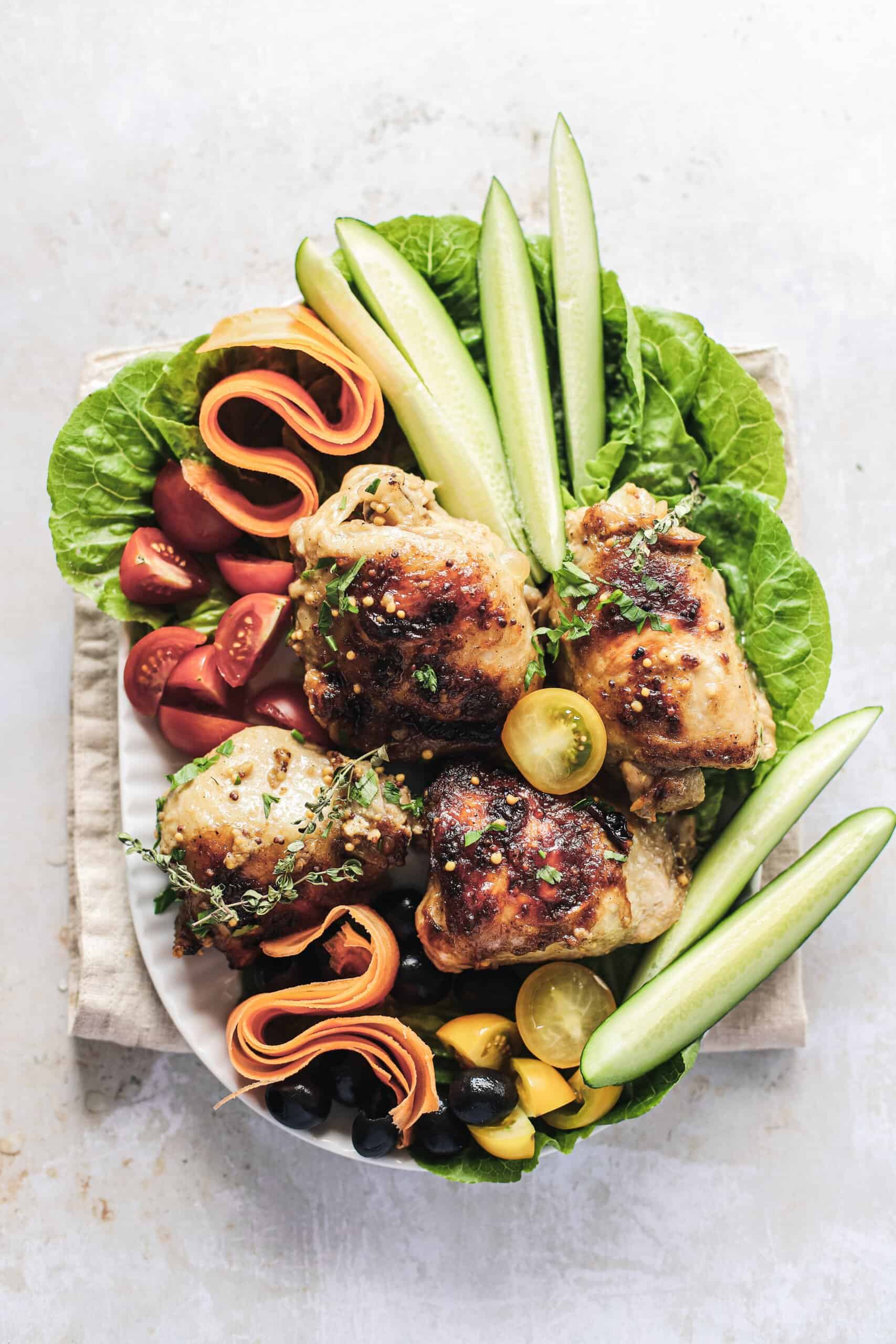 Four roasted chicken thighs on a serving platter with fruits and greens