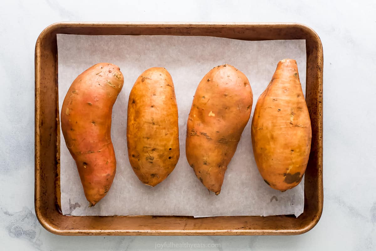 Four raw sweet potatoes sitting on a metal baking sheet lined with parchment paper