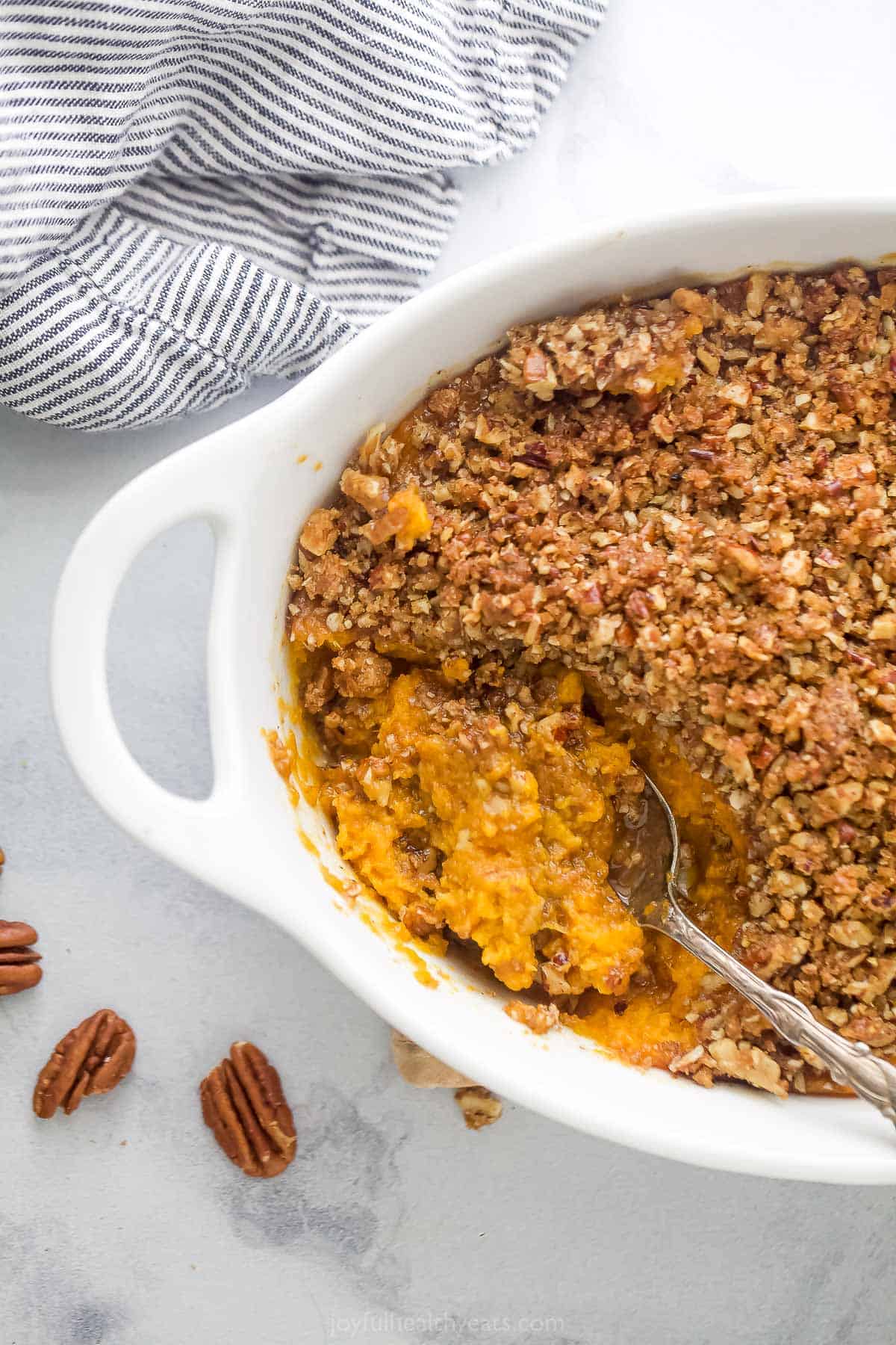 A white casserole dish containing a sweet potato casserole with a few pecans and a kitchen towel beside it