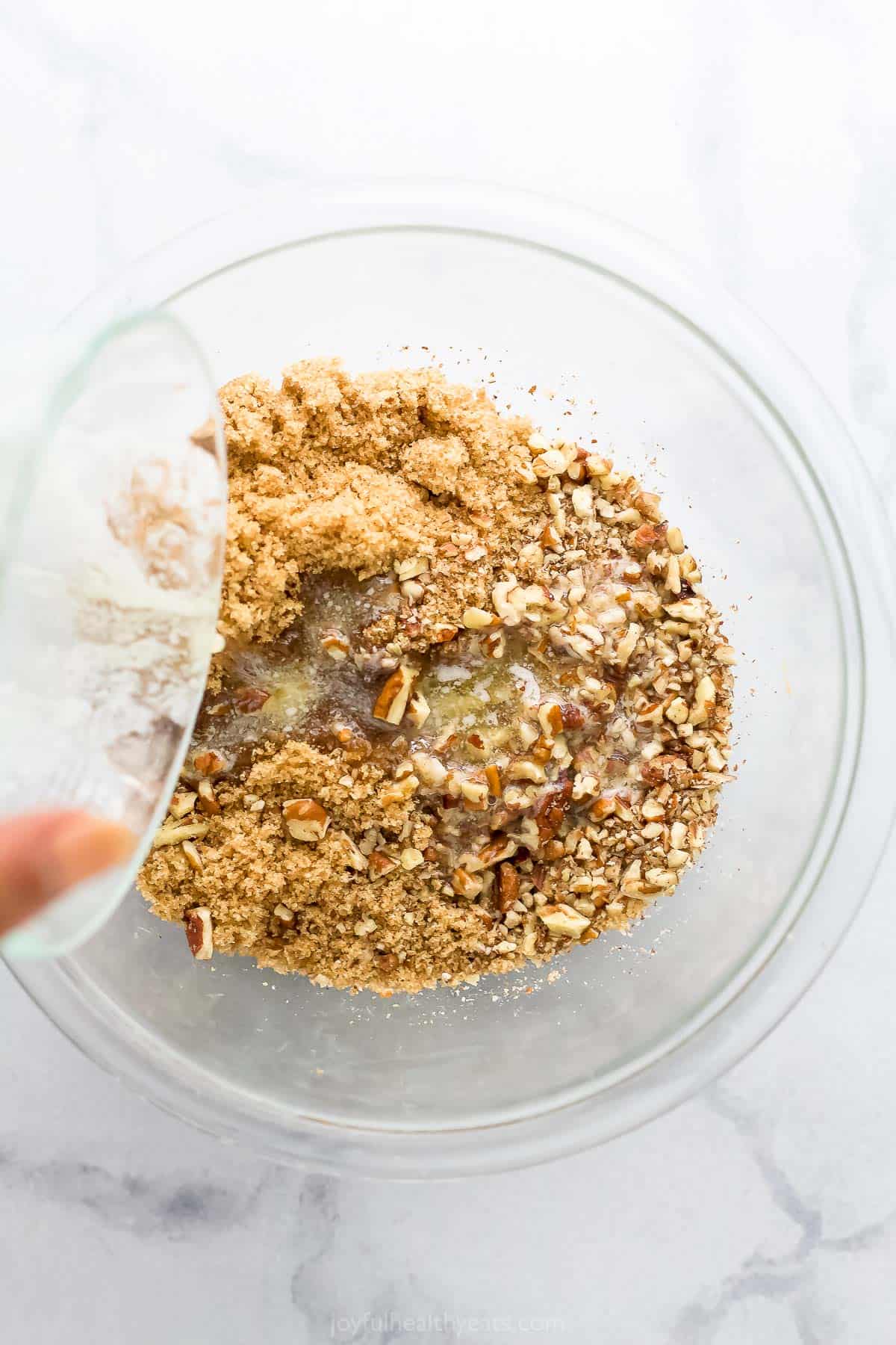 Melted butter being poured into a glass bowl containing chopped pecans, brown sugar and cinnamon