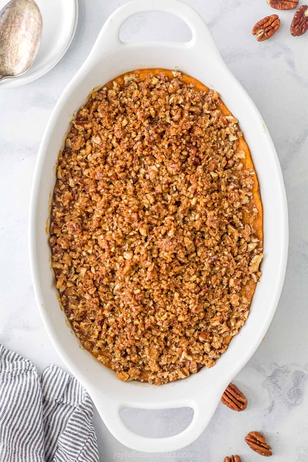 A baking dish with handles containing a freshly-baked sweet potato casserole with loose pecans on the counter beside it