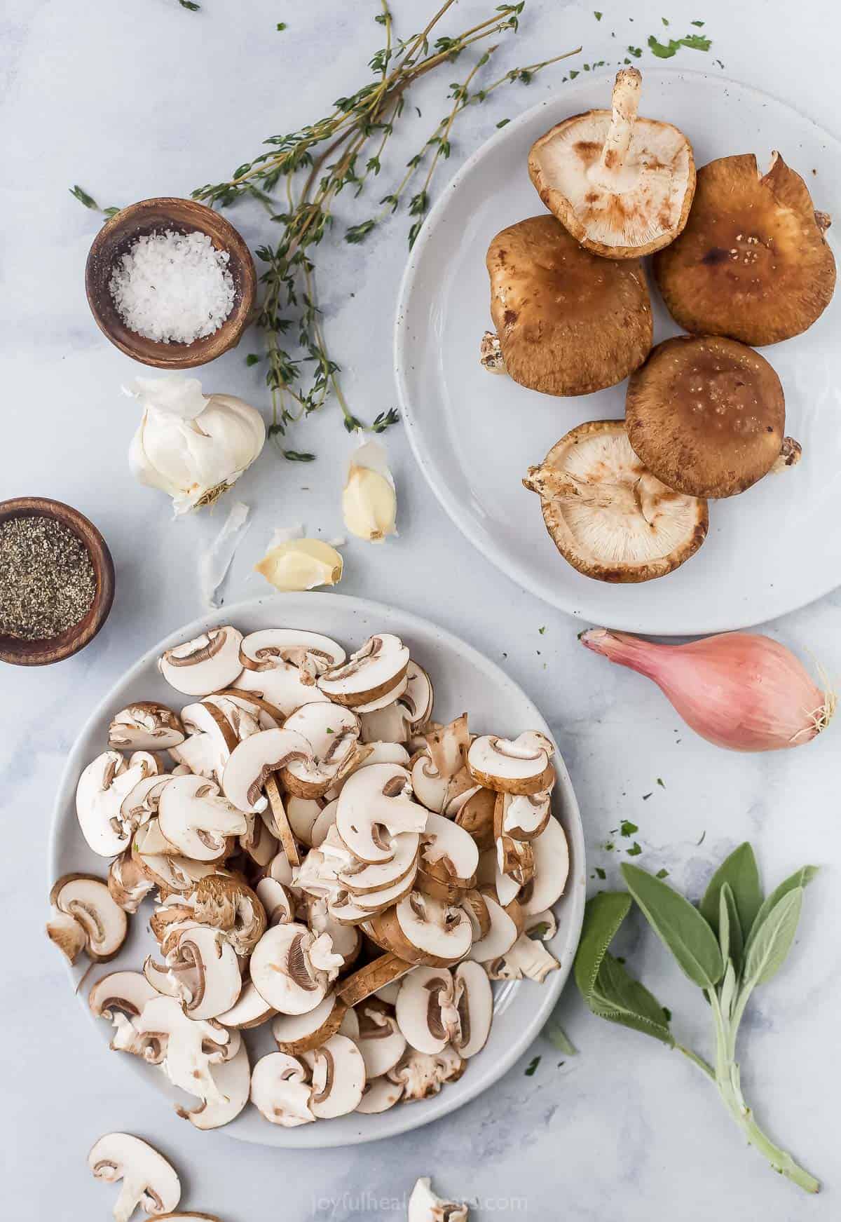 A plate of sliced mushrooms on a marble surface beside a plate of whole mushrooms and some loose garlic cloves