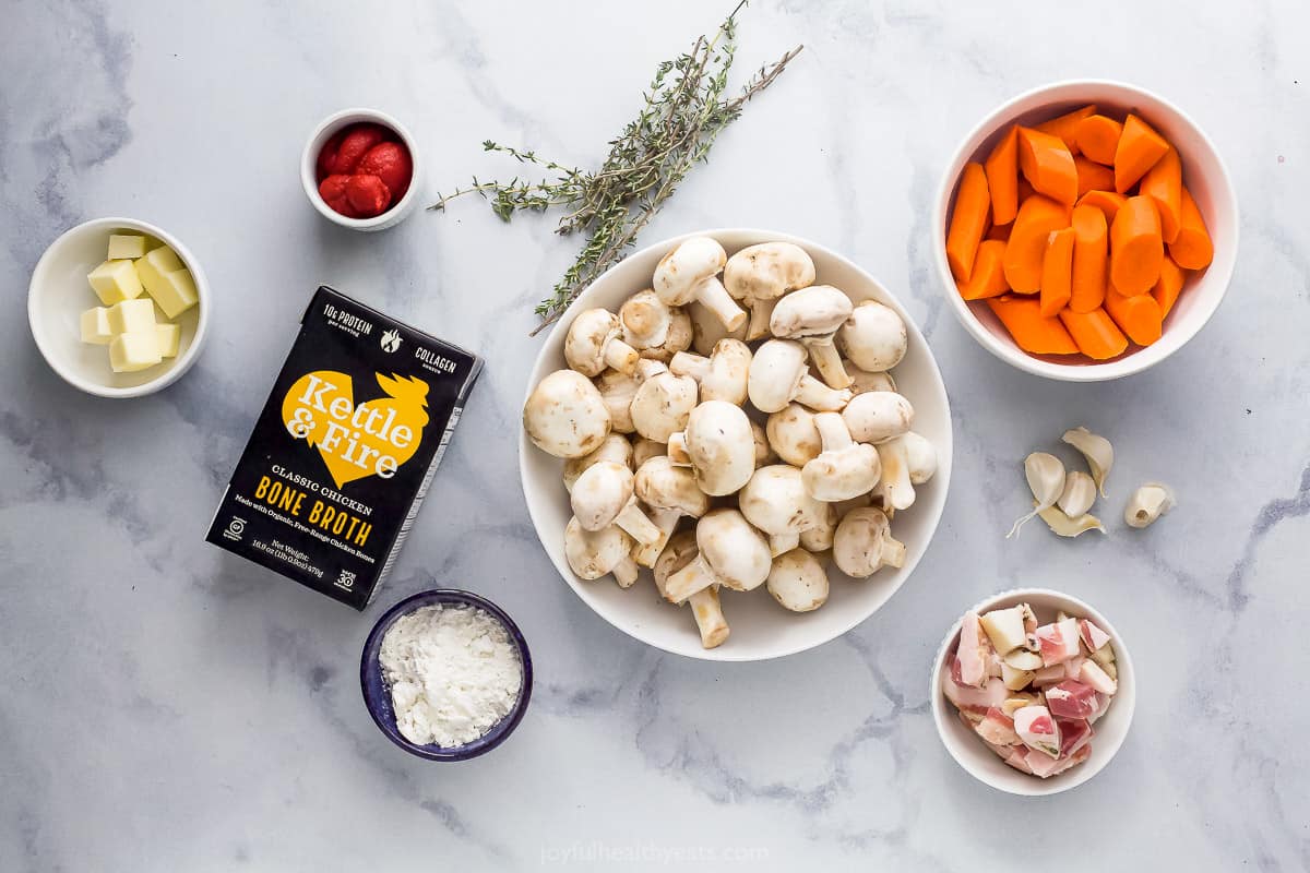 A bowl of mushrooms, a carton of bone broth, fresh thyme and many of the remaining stew ingredients on a marble countertop