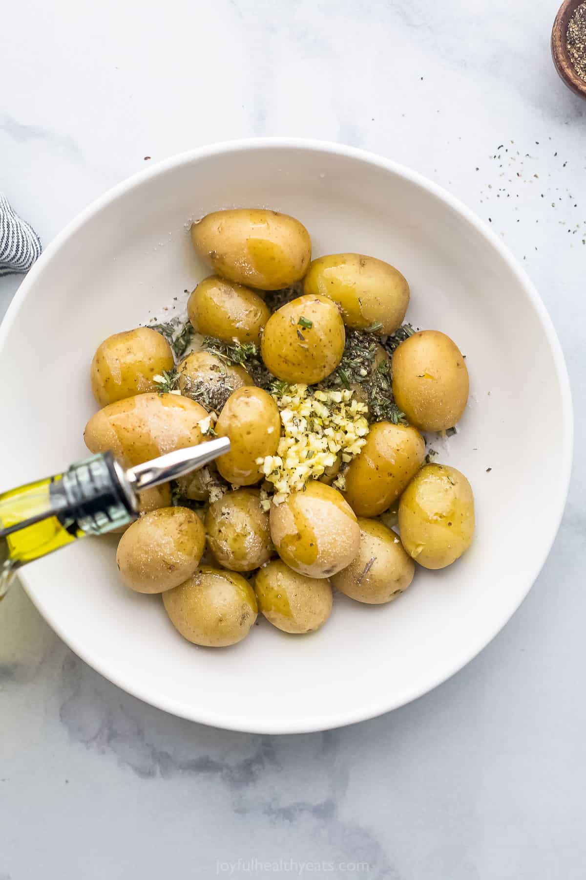Olive oil being poured into a bowl containing baby potatoes, minced garlic and chopped fresh herbs