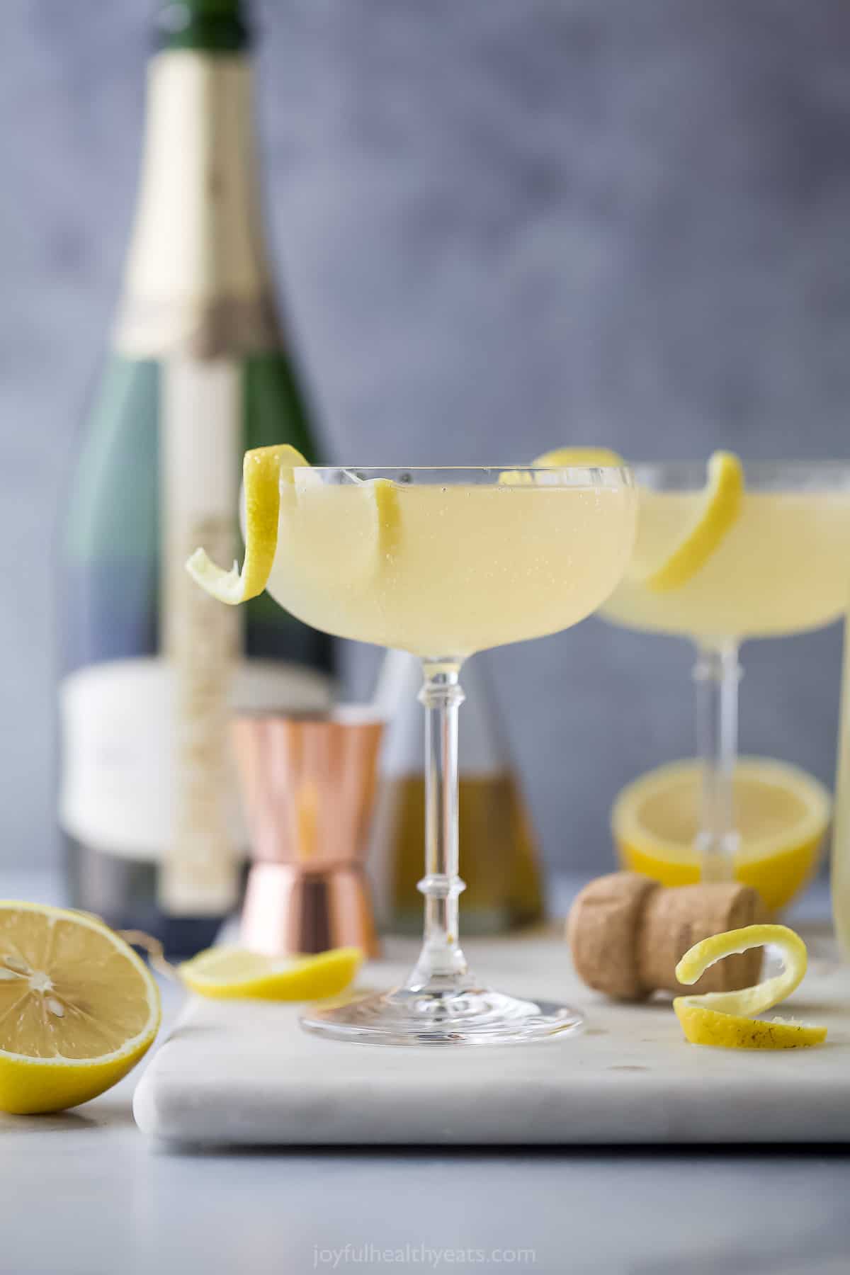 Two classic French 75 cocktails on a surface cluttered with a bottle of champagne and fresh lemon slices