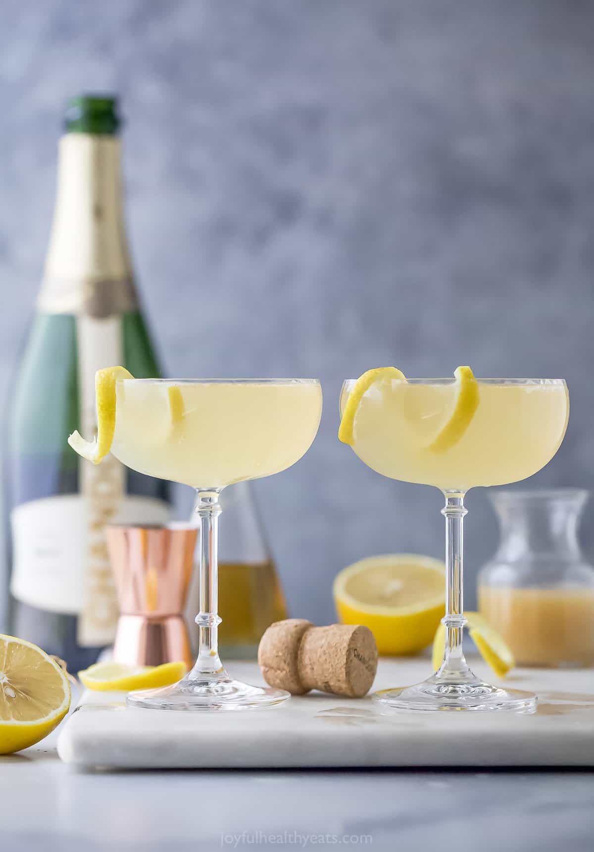 Two glasses of champagne mixed drinks on a cutting board with a cork and two lemon twists beside them