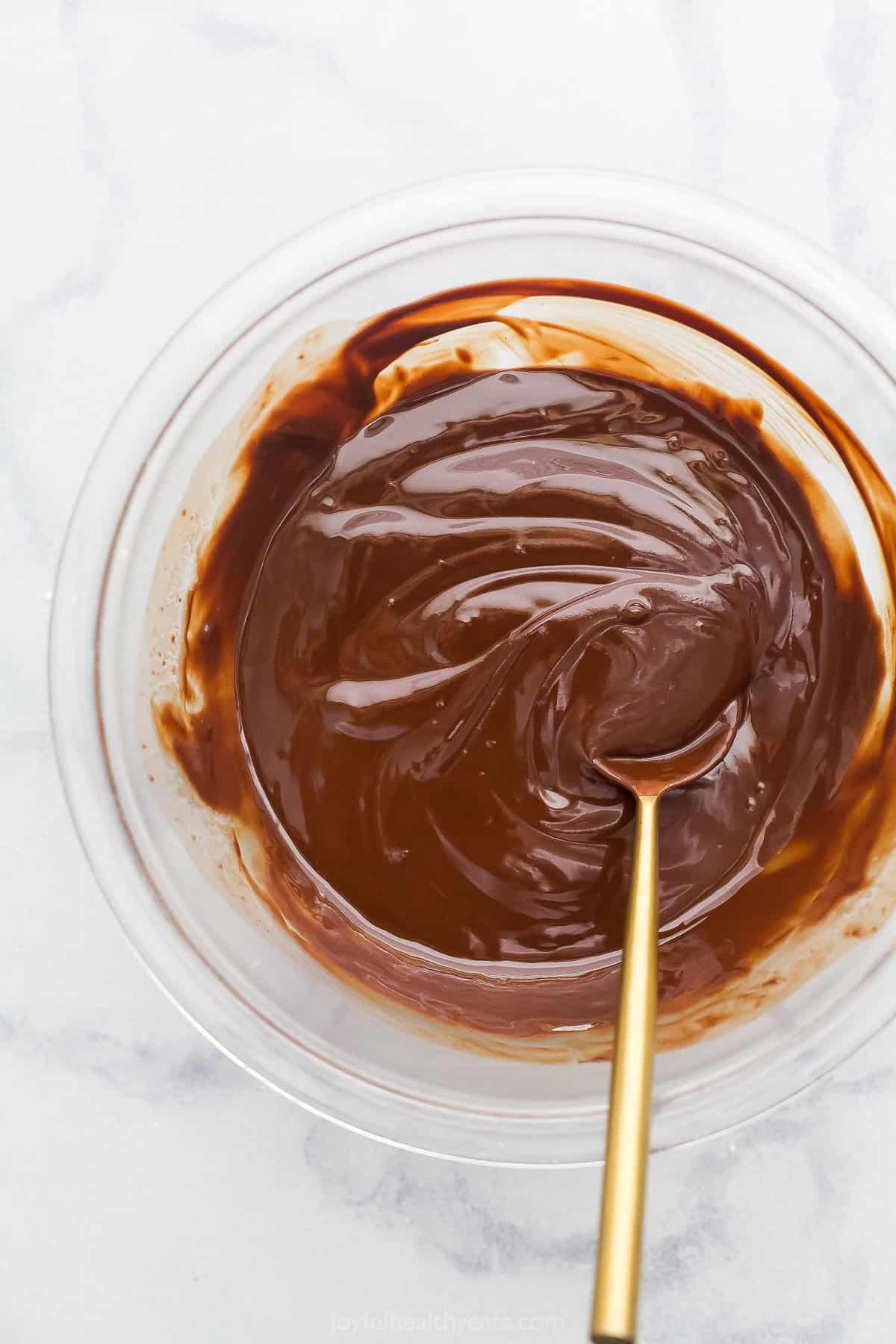 A bowl of melted chocolate and butter being stirred with a copper spoon