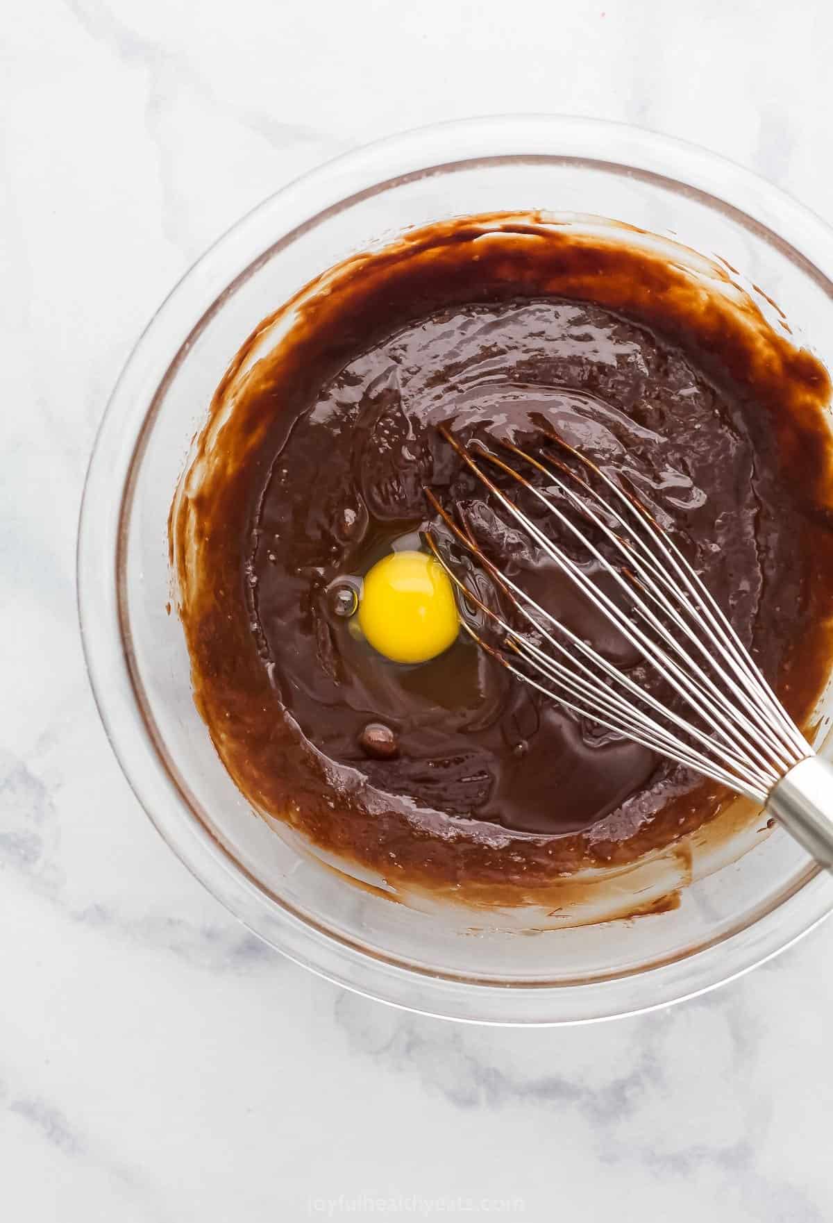 An egg being mixed into a bowl of melted chocolate with a large whisk