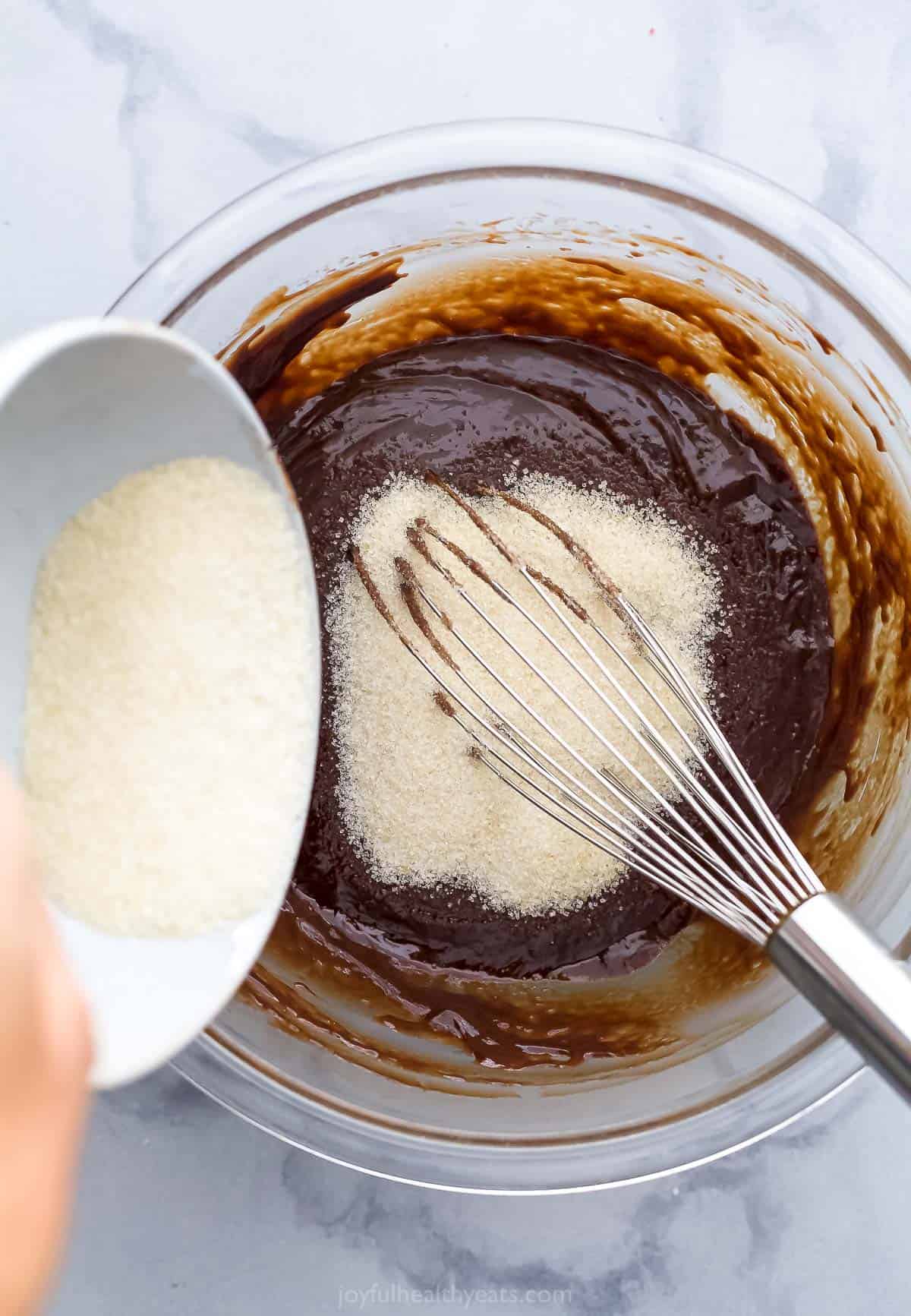 Sugar being poured into a large glass bowl containing brownie batter and a metal whisk
