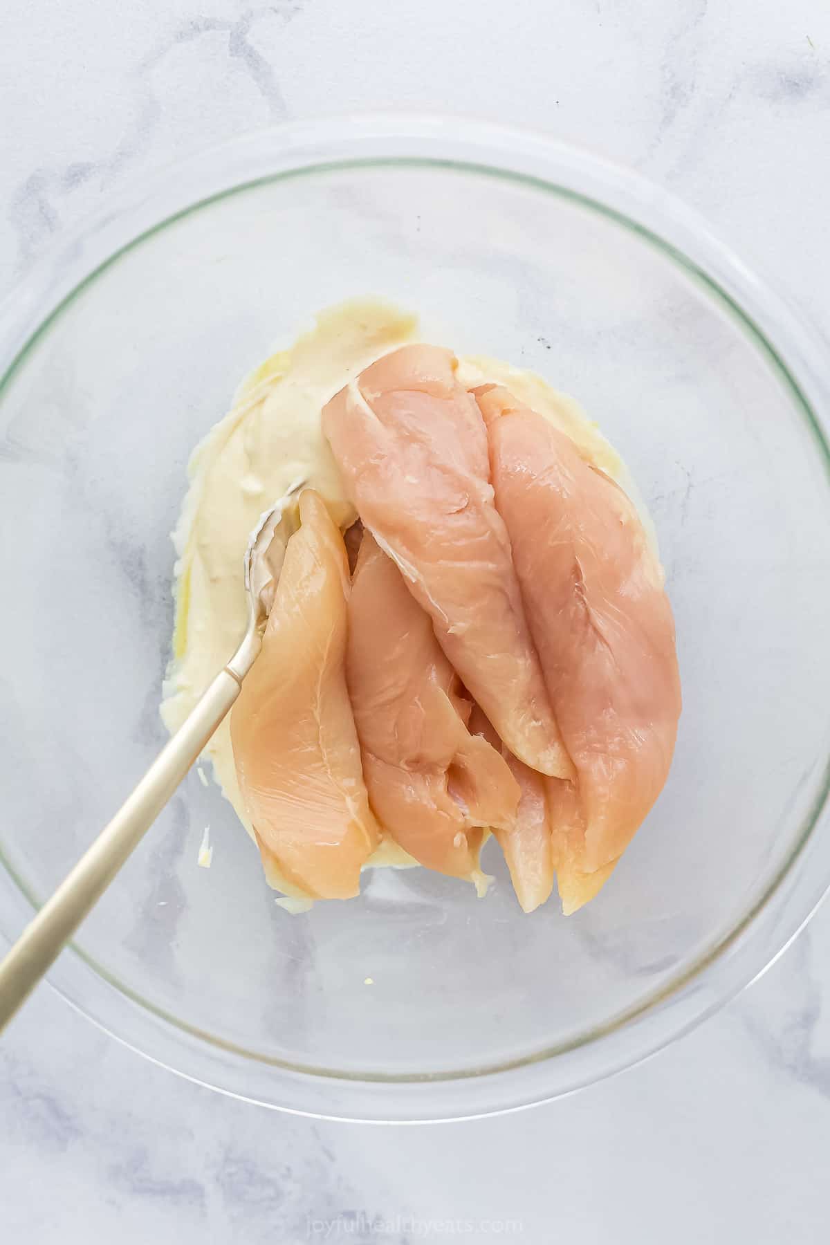 Four raw pieces of chicken in a glass mixing bowl with the mayonnaise mixture