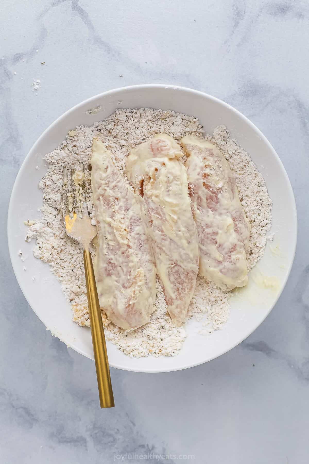 Three chicken tenders in a bowl with the combined dry ingredients and a golden fork