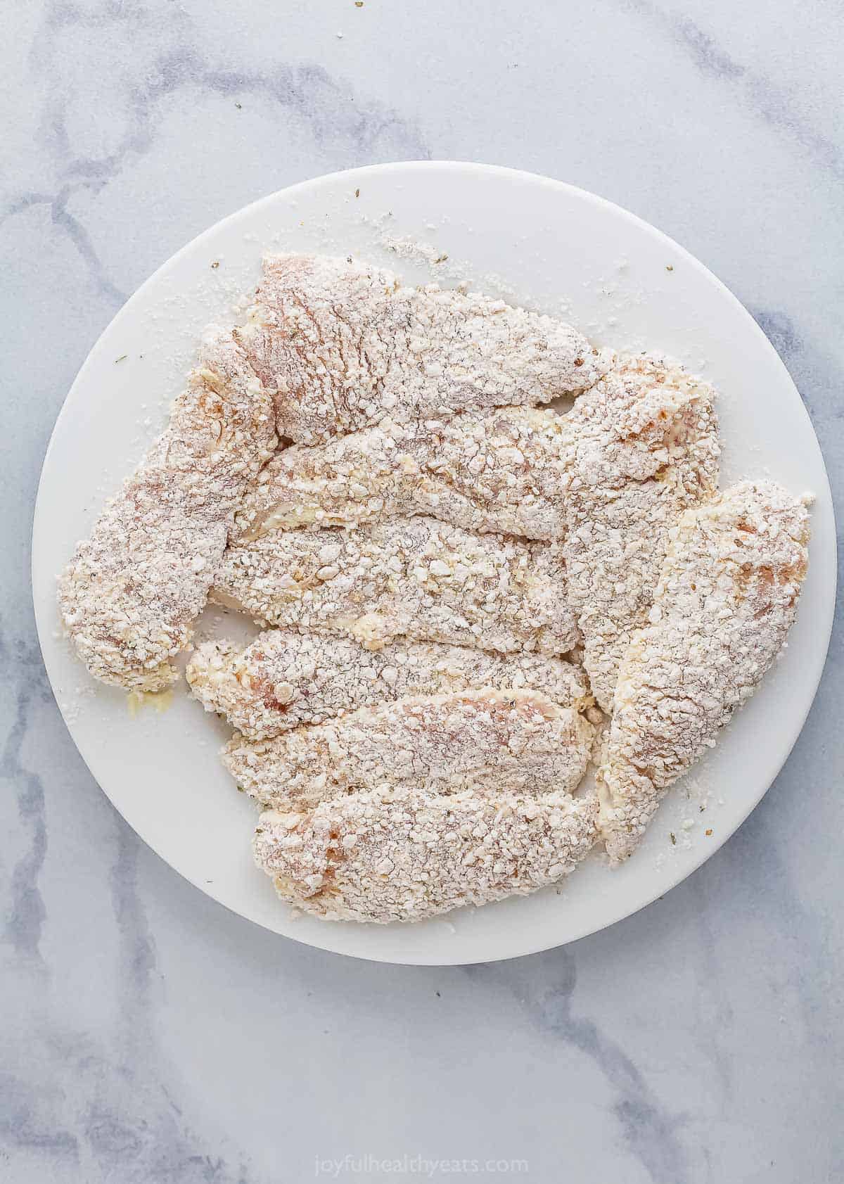 A plate of dredged chicken tenders sitting on a white and gray marble countertop