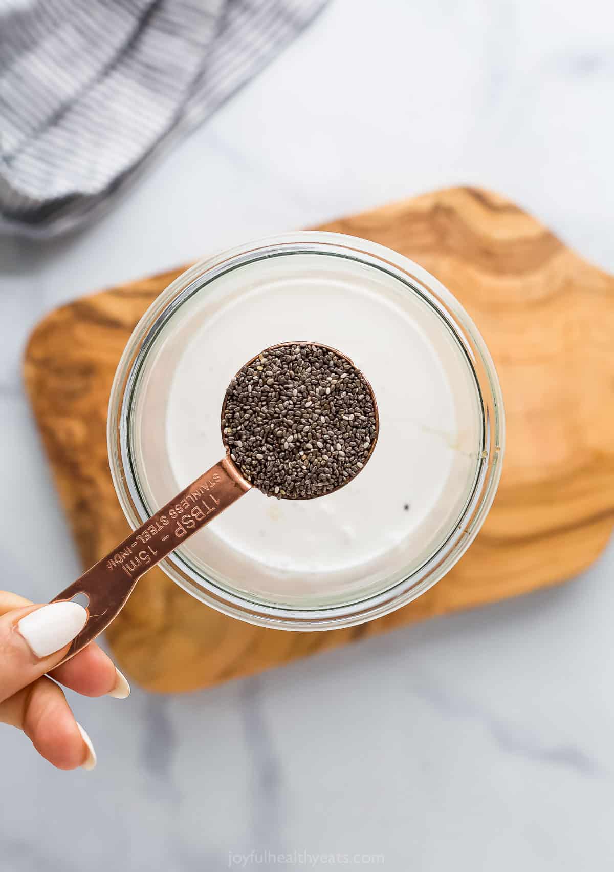 A tablespoon of chia seeds hovering over a jar filled with coconut cream, vanilla extract and maple syrup