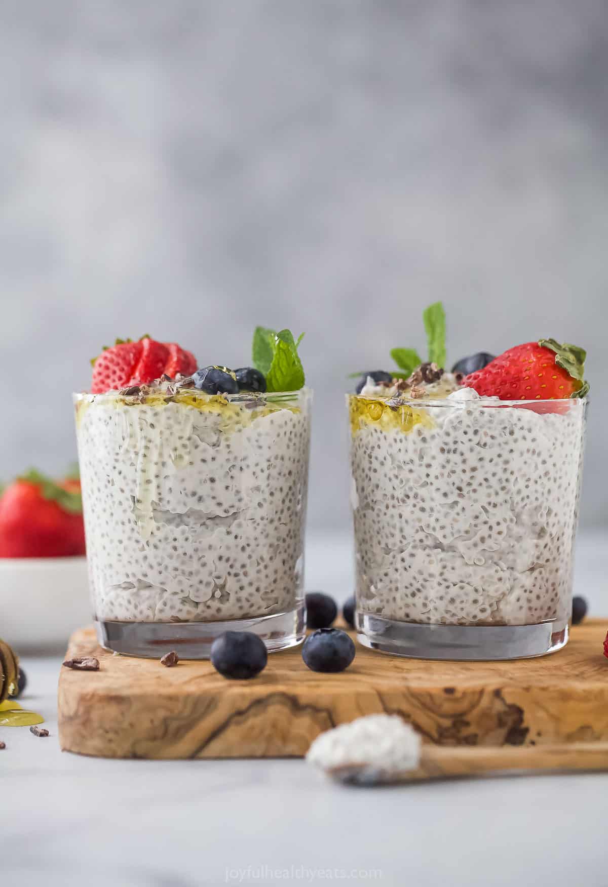 Two cups of chia seed pudding on a countertop with a bowl of fresh strawberries and a few loose blueberries