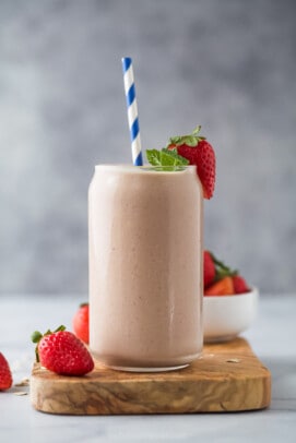 A homemade smoothie inside of a large glass with a striped straw inside and a fresh strawberry on the rim