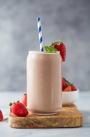 A homemade smoothie inside of a large glass with a striped straw inside and a fresh strawberry on the rim
