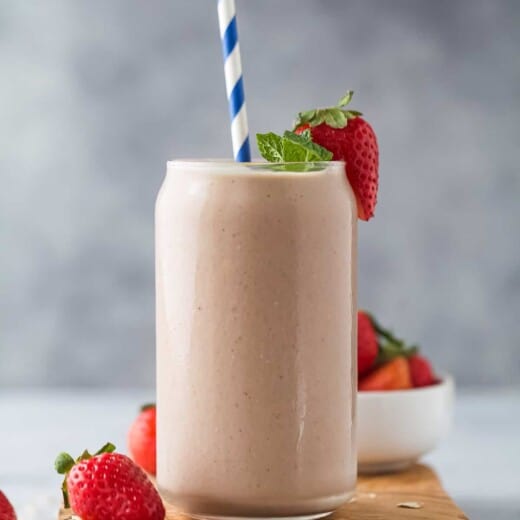 A homemade smoothie inside of a large glass with a striped straw inside and a fresh strawberry on the rim