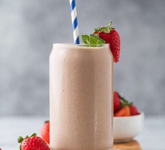 A homemade smoothie inside of a large glass with a striped straw inside and a fresh strawberry on the rim