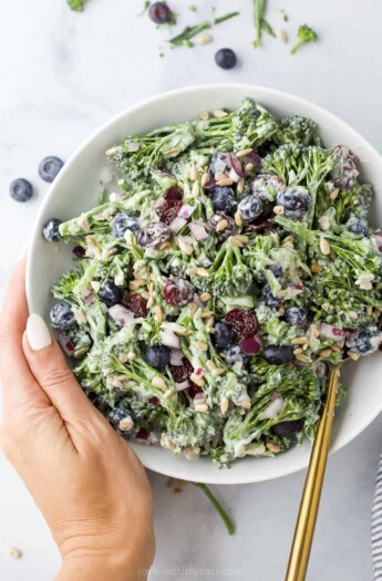 A hand holding a bowl of broccolini salad with fresh blueberries, sunflower seeds and dried cherries