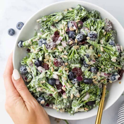 A hand holding a bowl of broccolini salad with fresh blueberries, sunflower seeds and dried cherries