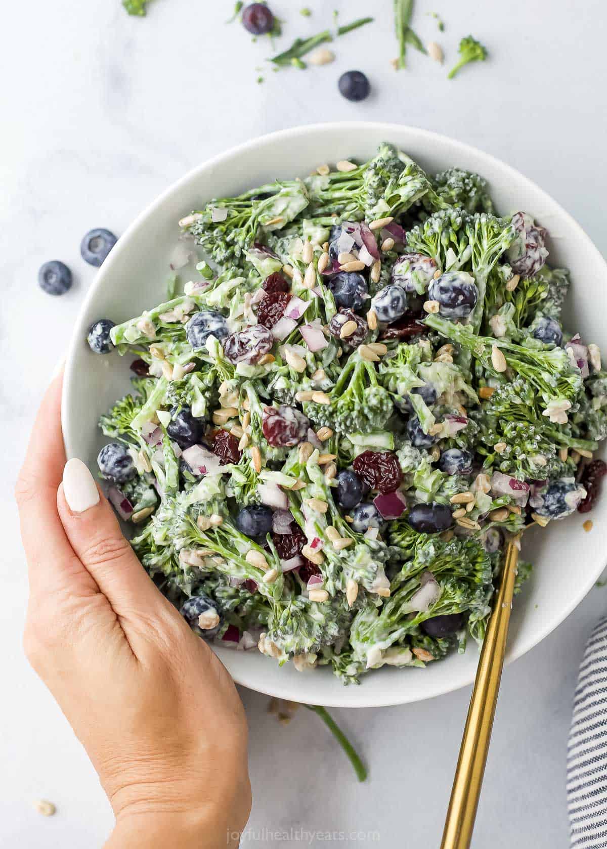 A hand holding a bowl of broccolini salad with fresh blueberries, sunflower seeds and dried cherries