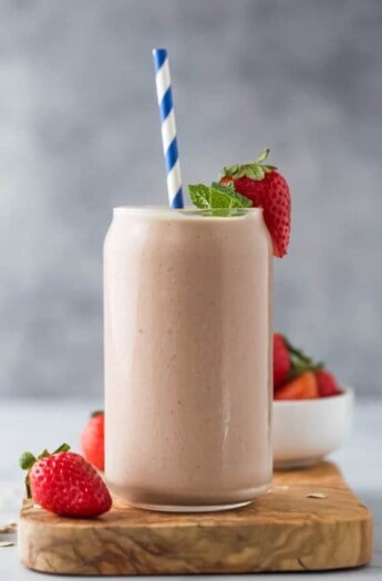 A homemade smoothie inside of a large glass with a striped straw inside and a fresh strawberry on the rim