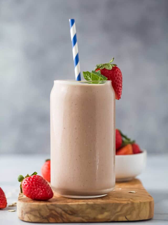 a pale pink smoothie in a glass with a straw on a wooden cutting board