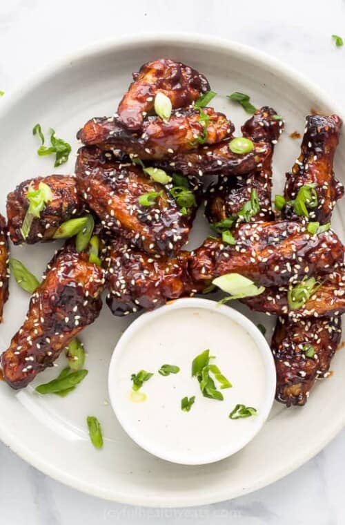 Overhead view of Asian chicken wings on a plate garnished with sesame seeds and green onion, next to a bowl of dressing.