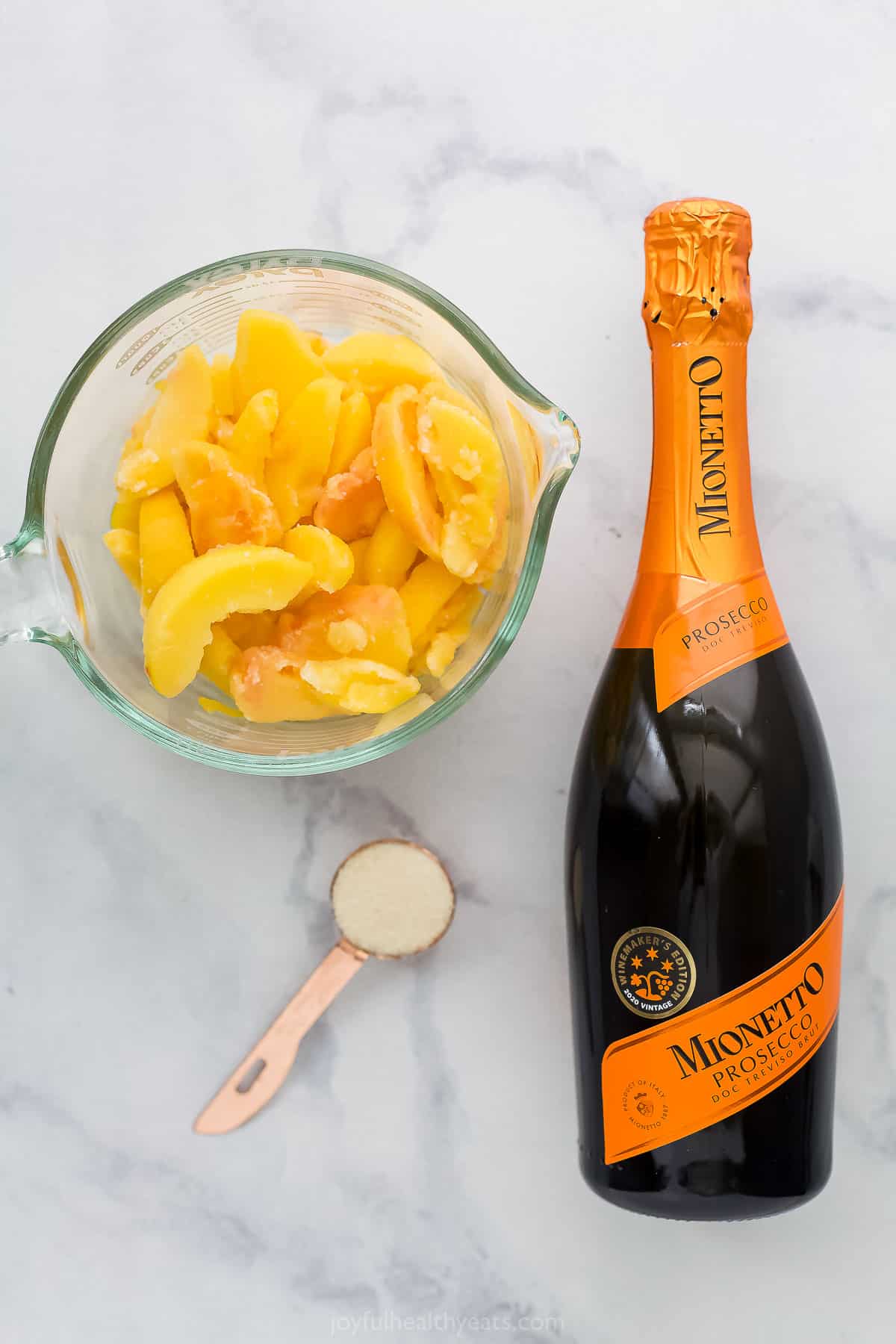 A bottle of prosecco on a marble countertop with sliced peaches and granulated sugar