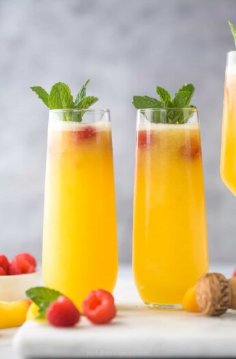 Two peach bellinis beside each other on a gray cutting board with a cork and two fresh raspberries