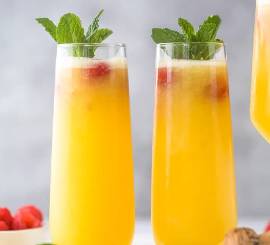Two peach bellinis beside each other on a gray cutting board with a cork and two fresh raspberries