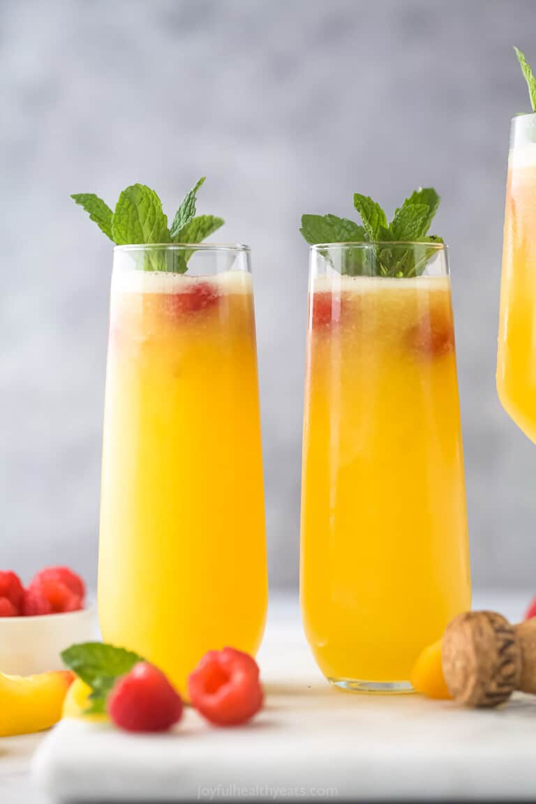Two peach bellinis beside each other on a gray cutting board with a cork and two fresh raspberries