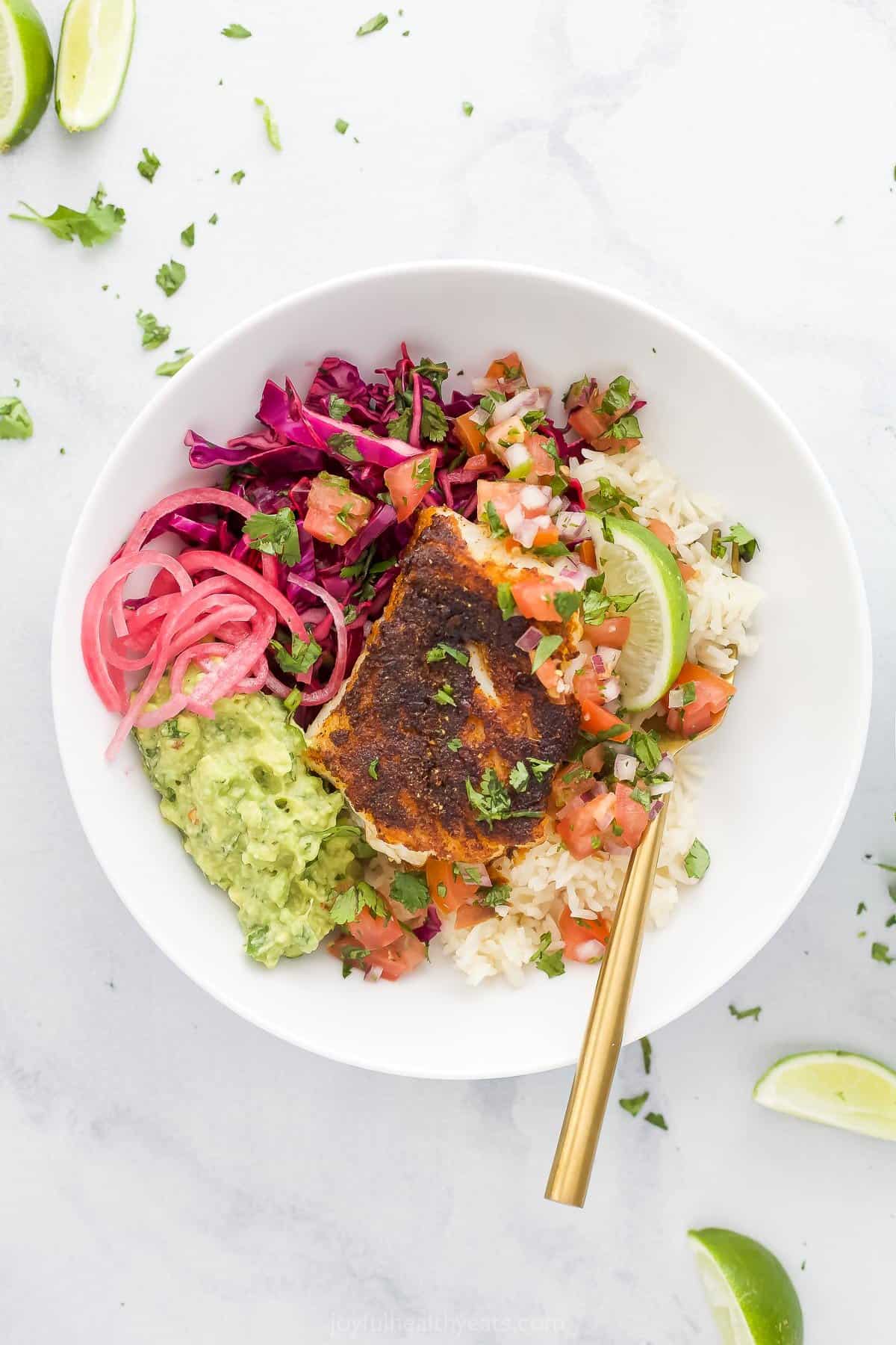 The bird's-eye view of a fish taco rice bowl on a marble surface beside four lime wedges