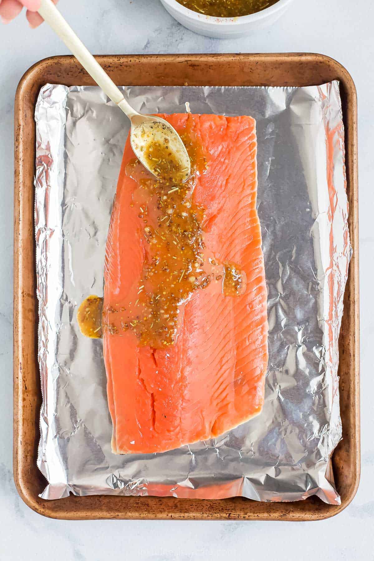 A raw fish fillet on a lined baking sheet with homemade glaze being spooned over it