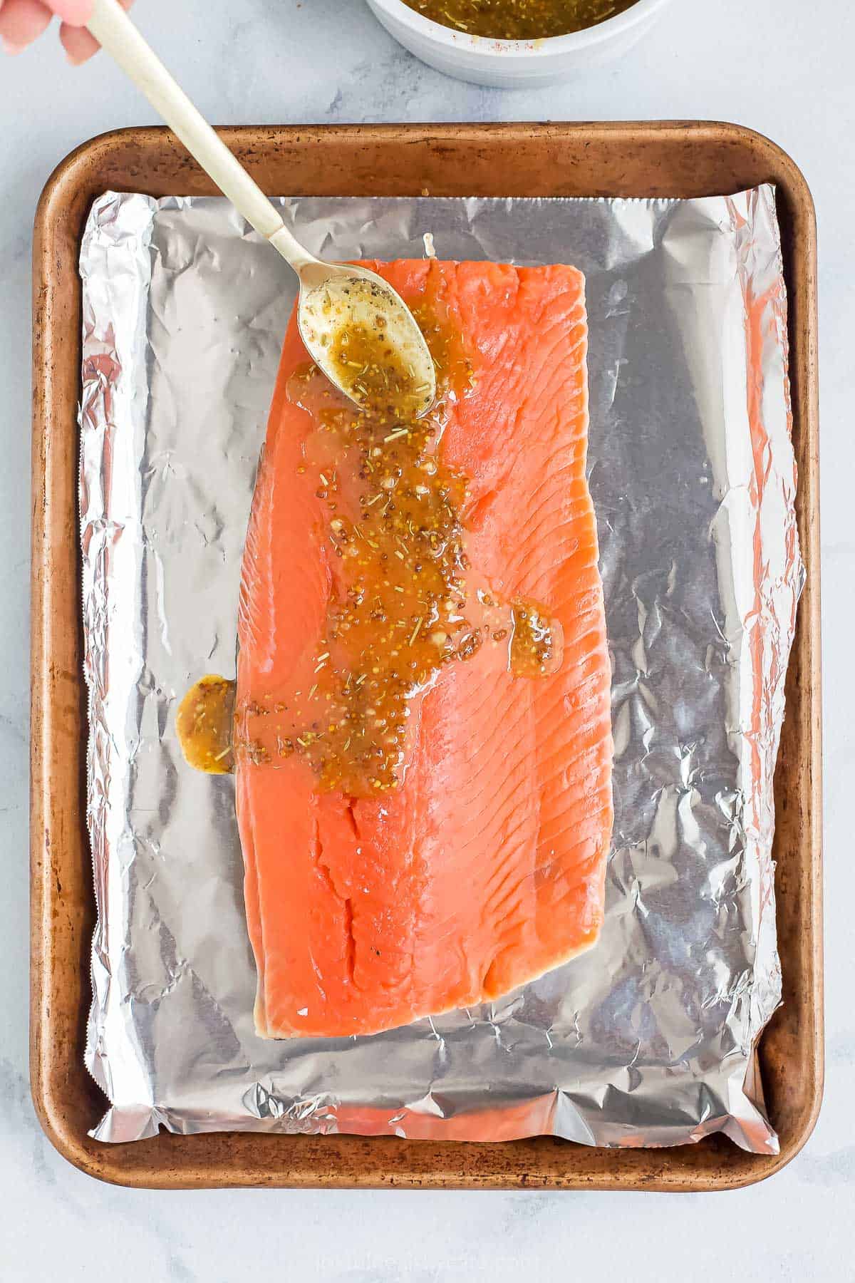 A raw fish fillet on a lined baking sheet with homemade glaze being spooned over it.